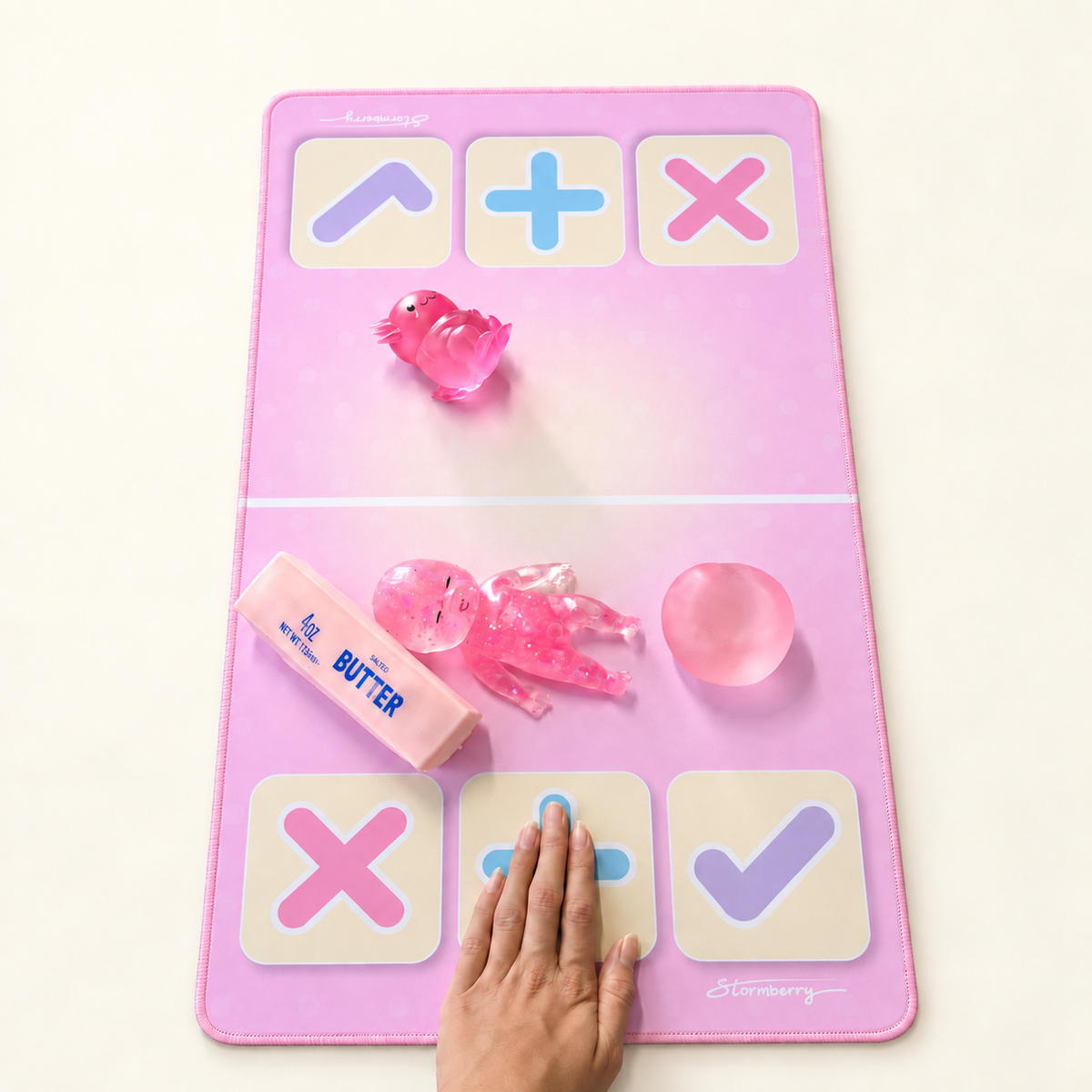 A hand taps the blue plus symbol on a Signature Pink trading mat with squishies on opposite sides of the divider.